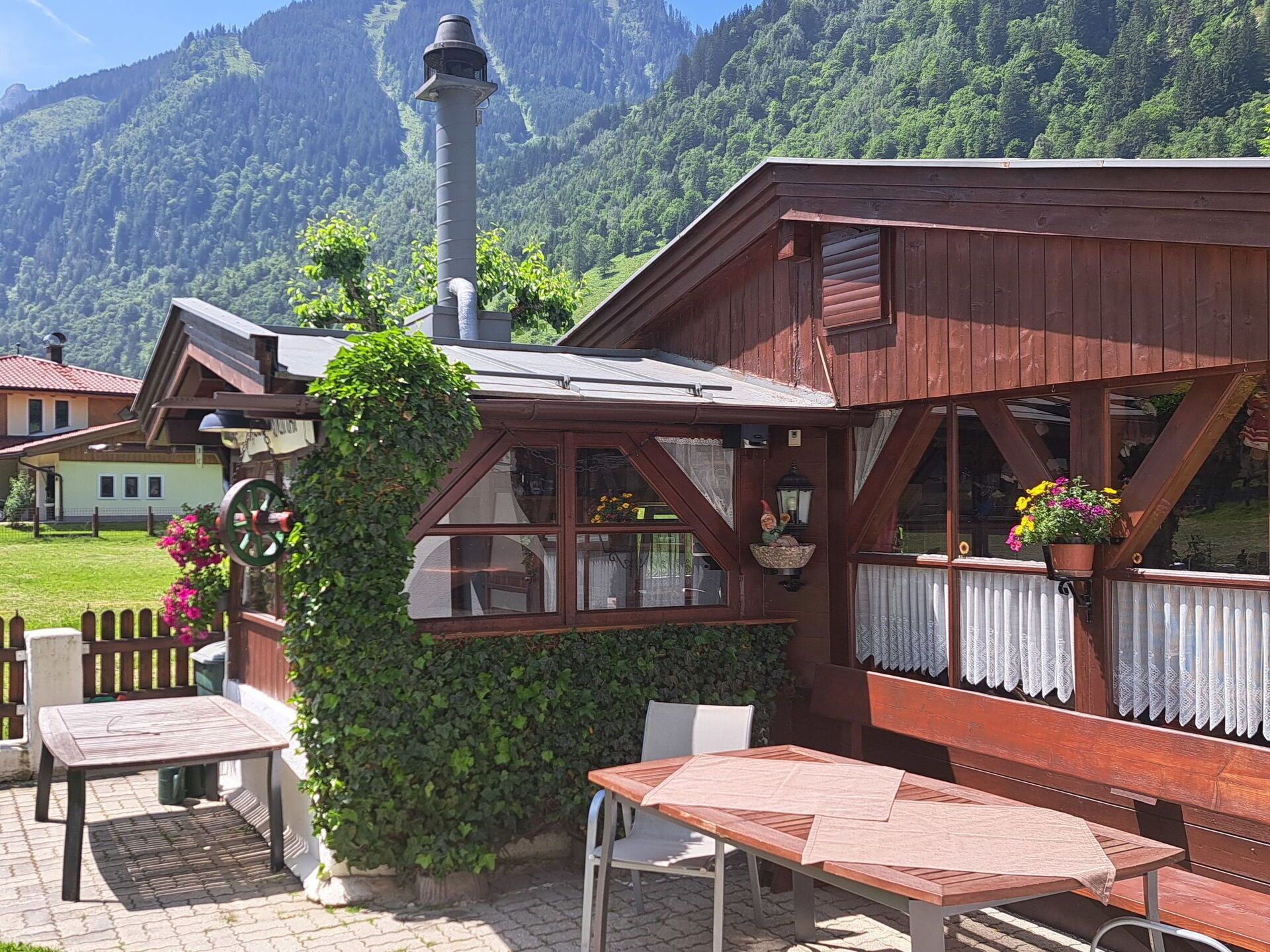 A rustic wooden building with a chimney, a garden area with a table and chairs, and a mountain backdrop.