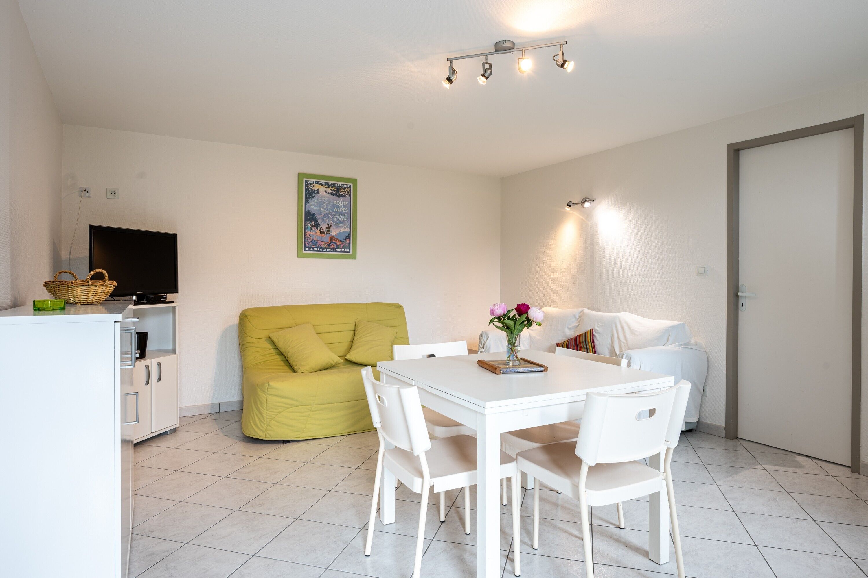Une pièce moderne et bien éclairée, comprenant une table à manger, des chaises, un canapé, un téléviseur et une affiche accrochée au mur.