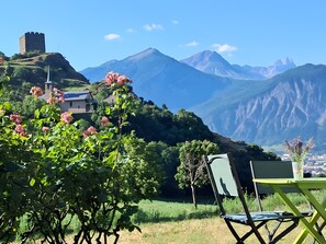 A mountainous landscape with a castle on a hill, lush greenery, and a table with a chair.