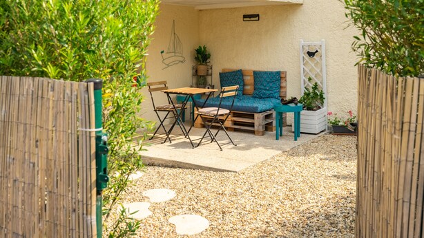 Une terrasse avec une table et des chaises en bois, un coin salon bleu avec des coussins, et un mur blanc orné d’un dessin de voilier.