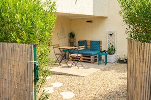 Un patio con una mesa y sillas de madera, un área de descanso con cojines azules y una pared blanca decorada con un dibujo de un velero.