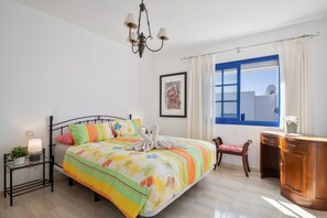 A bedroom with a bed, bedside table, lamp, chair, desk, window, and artwork.