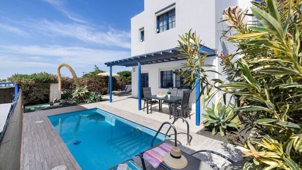 A swimming pool with a wooden deck, a patio area with a table and chairs, and a white building with blue accents.