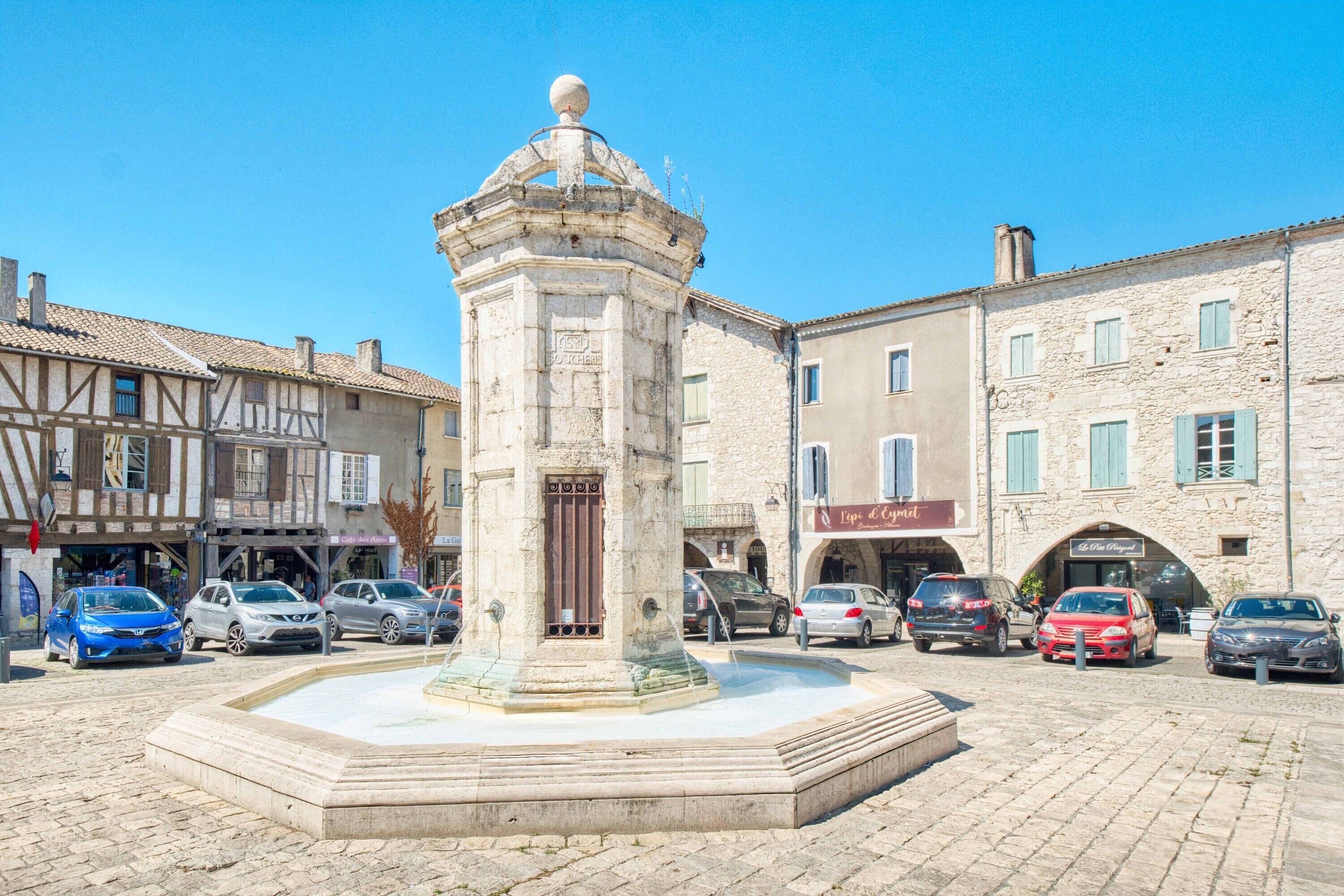 Una plaza con una fuente central, rodeada de edificios de piedra con tiendas y coches aparcados.