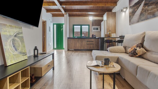 A modern living room with a wooden ceiling, a flat-screen TV, a sofa, a coffee table, and a kitchen in the background.