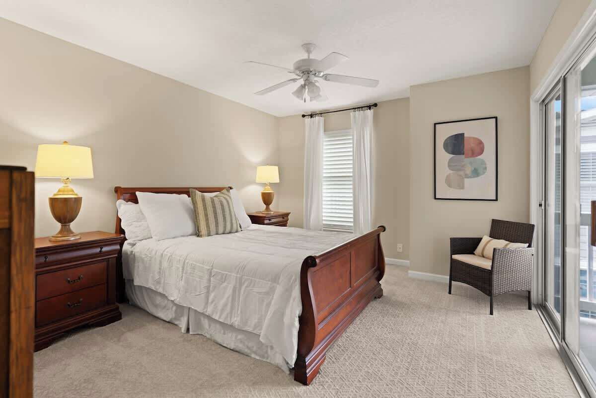 Une chambre à coucher avec un lit, deux tables de chevet avec des lampes, une chaise, un ventilateur de plafond et une fenêtre avec des stores.
