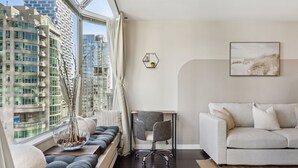 A modern living room with a sofa, a desk, and a chair. A large window offers a view of tall buildings.
