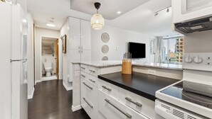 A modern kitchen with white cabinetry, a black countertop, and a built-in oven. There is a hanging light fixture and a window providing a view of the city.
