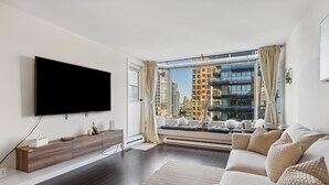 A modern living room with a large flat-screen TV mounted on the wall, a sofa with decorative pillows, a wooden console table, and a large window offering a city view.