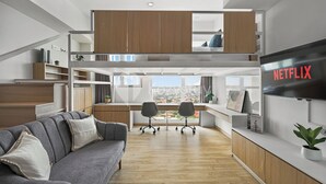 Un appartement moderne de style « loft », doté d’un lit en mezzanine, d’un coin repas, d’un espace de travail avec un bureau et des chaises, d’un canapé gris et d’un grand téléviseur.