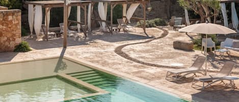 A pool area with a clear water section, lounge chairs, and a pergola with draped fabric.