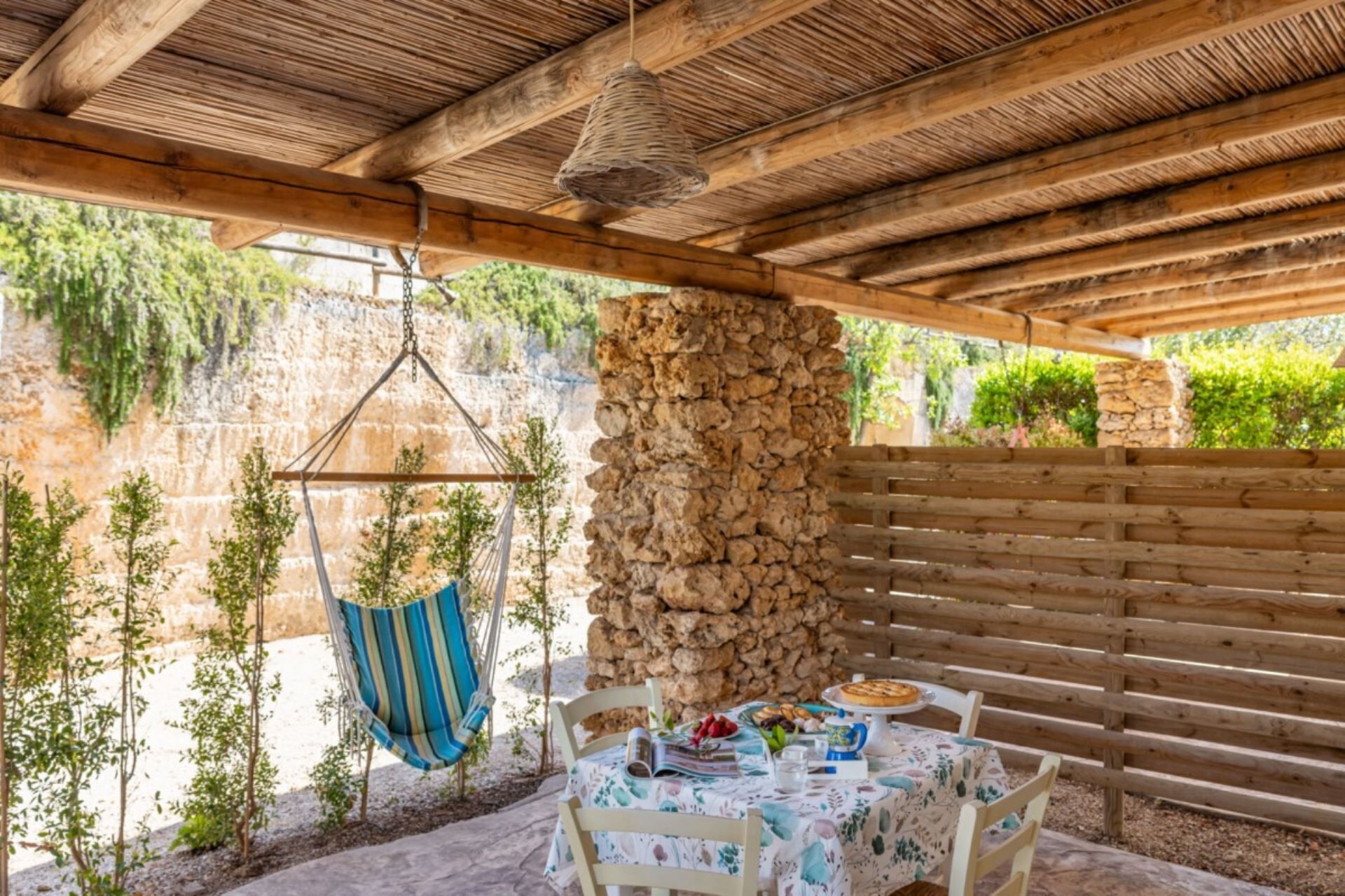 An outdoor patio with a stone wall, wooden bench, hanging chair, and a table set with food.