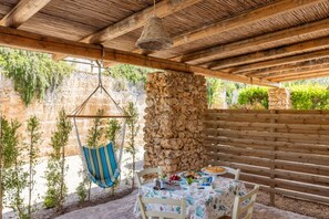 An outdoor patio with a stone wall, wooden bench, hanging chair, and a table set with food.