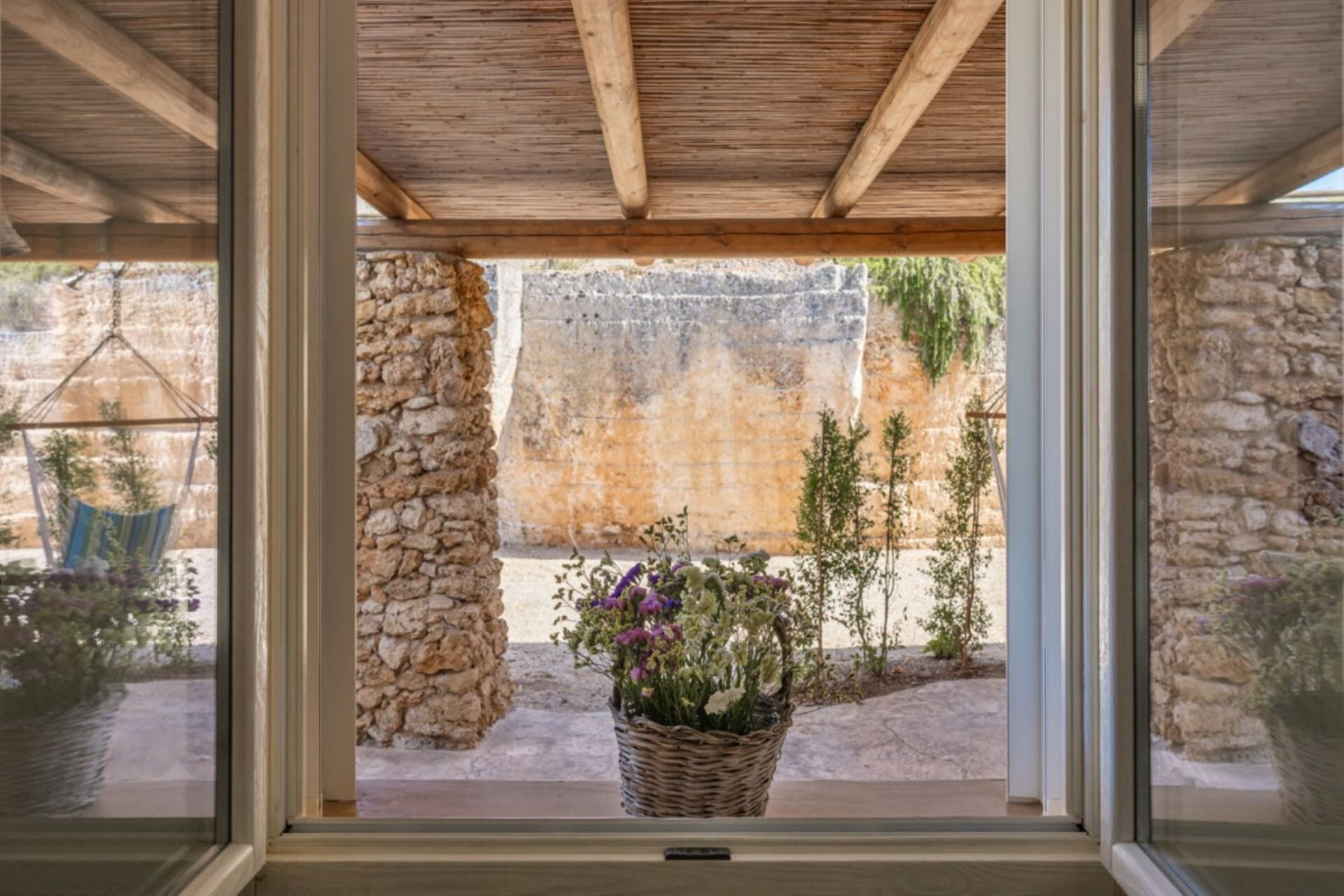 A view through a window with a wooden ceiling and stone walls, a hanging chair, and a basket of flowers.