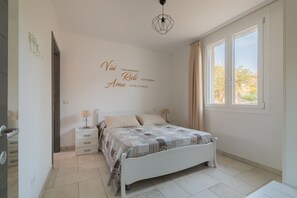 Quarto com cama, duas mesas de cabeceira, parede com frase motivacional, janela com cortinas e luminária no teto.