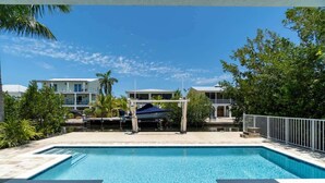 A rectangular swimming pool with a clear blue water surface, surrounded by a stone deck. In the background, there are two-story buildings with balconies and a covered area with a blue roof.