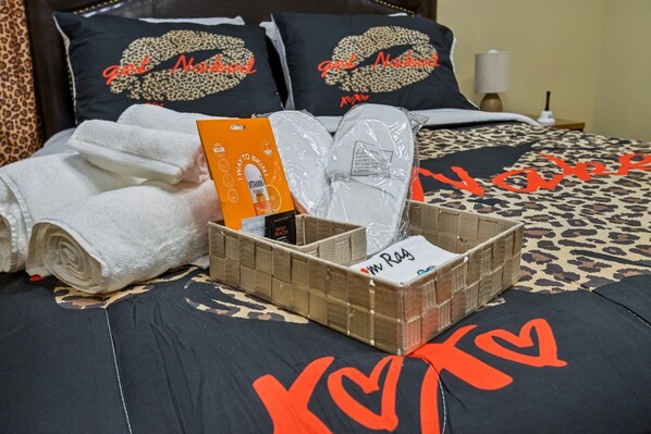 A neatly made bed with a leopard print bedspread and pillows with a heart design. There are rolled white towels, a basket with toiletries, and a box of lotion.