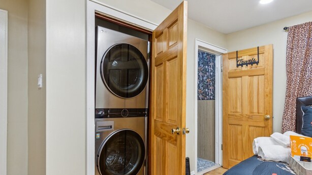 A laundry room with two stacked washing machines, a wooden door, a towel rack, and a bed with a blue blanket.