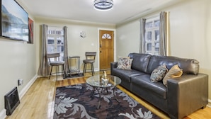 A living room with a leather sofa, a glass coffee table, and a patterned rug. There are two chairs, a door, and a window with curtains.