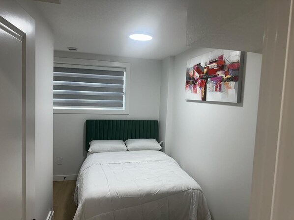 A small, neatly arranged bedroom with a single bed, a window with blinds, and a colorful abstract painting on the wall.