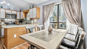 A modern kitchen with a dining area, featuring a central island, wooden cabinets, and a large window with curtains. - 3BR Turtle Hut - Beach House w/ Fenced Yard & Firepit (Jacksonville Beach)
