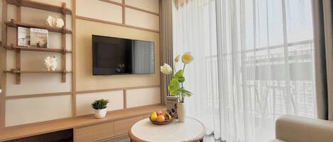 A modern living room with a flat-screen TV mounted on the wall, a round coffee table with fruit and a vase, a sofa, and a large window with sheer curtains.