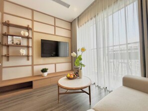 A modern living room with a flat-screen TV mounted on the wall, a round coffee table with fruit and a vase, a sofa, and a large window with sheer curtains.