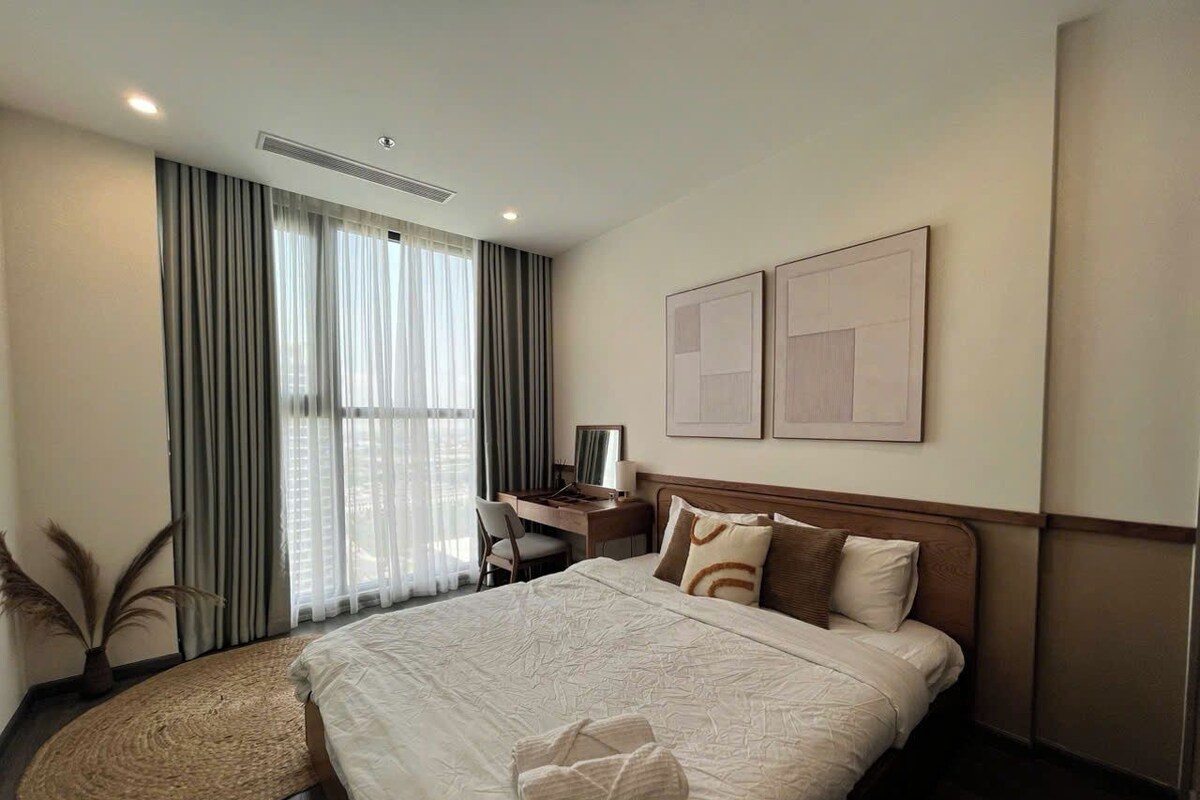A hotel room with a large bed, a desk, and a chair. There are two paintings on the wall and a window with curtains.
