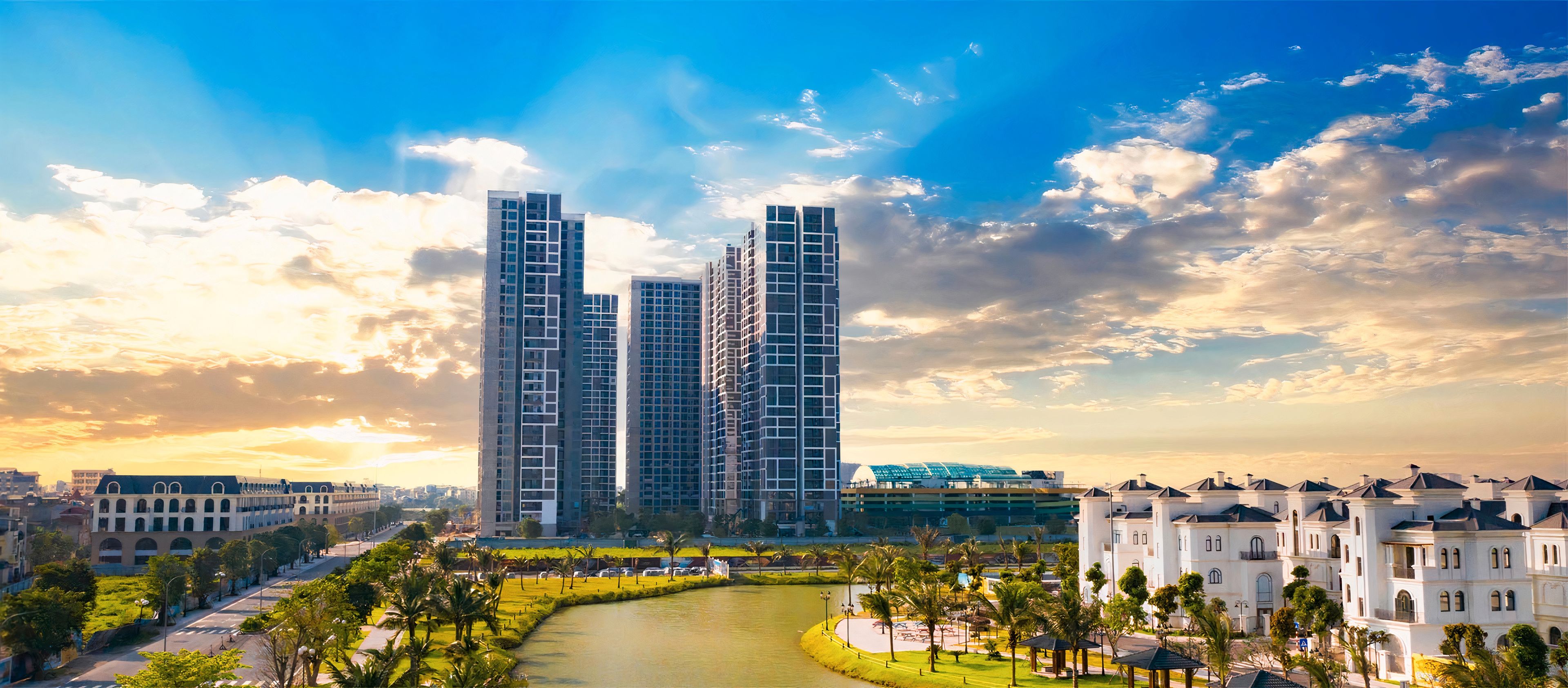 A modern high-rise building complex with a river in the foreground and a park with palm trees.