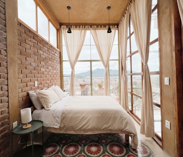 Un dormitorio con pared de ladrillo, cama con ropa de cama blanca, una mesita con lámpara y ventanas grandes con cortinas.