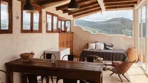 A rustic room with a wooden ceiling, a bed with pillows, a dining table with chairs, and a view of a mountainous landscape through large windows.