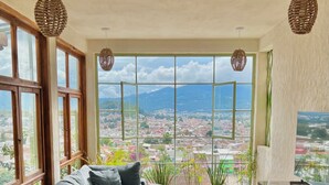 A modern living room with a large sectional sofa, a small round table, and a TV stand. The room has large windows offering a city view.