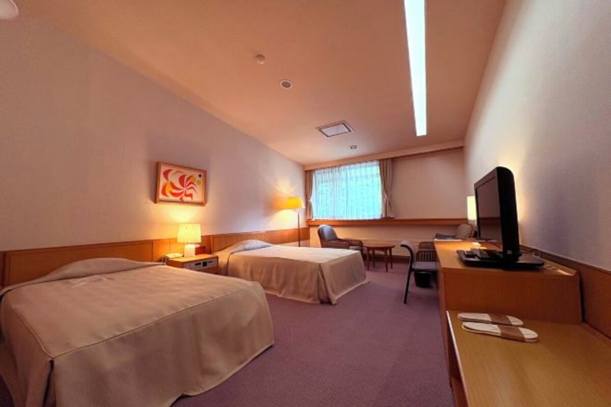 A hotel room with two beds, a desk, a chair, a TV, and a window with curtains.