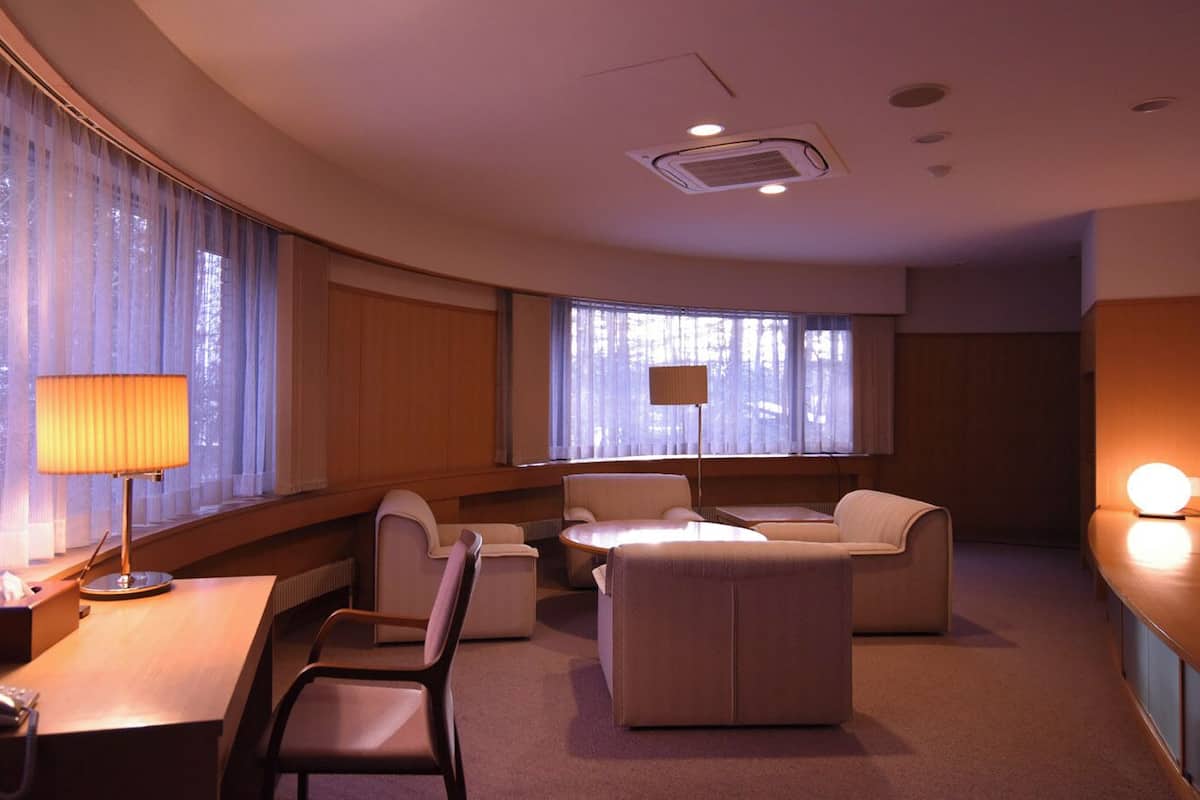 A hotel room with a desk, two chairs, a sofa, a TV, and a lamp.