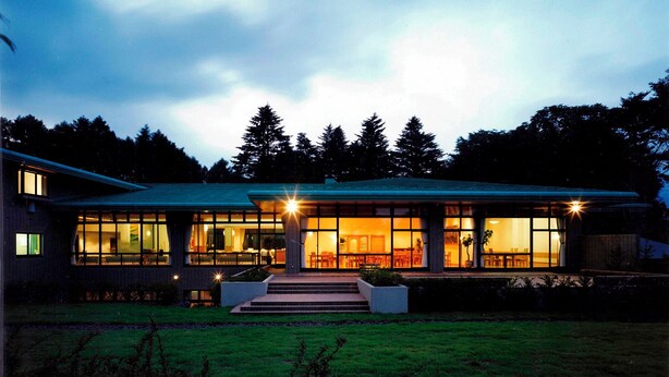 A modern house with large windows and a well-lit interior, surrounded by a grassy yard and trees.