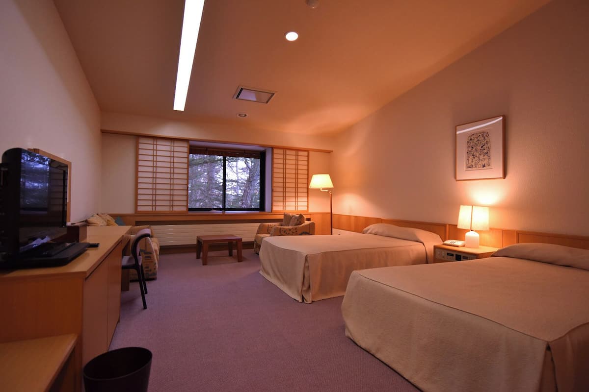 A hotel room with two beds, a desk with a computer, a chair, a window with blinds, a lamp, and a framed picture on the wall.