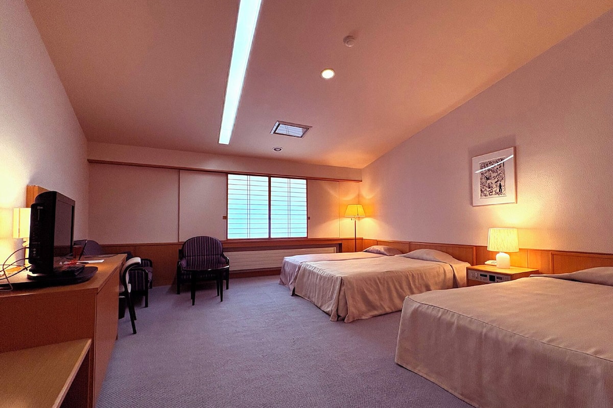 A hotel room with two beds, a desk with a computer, a chair, and a window with blinds.