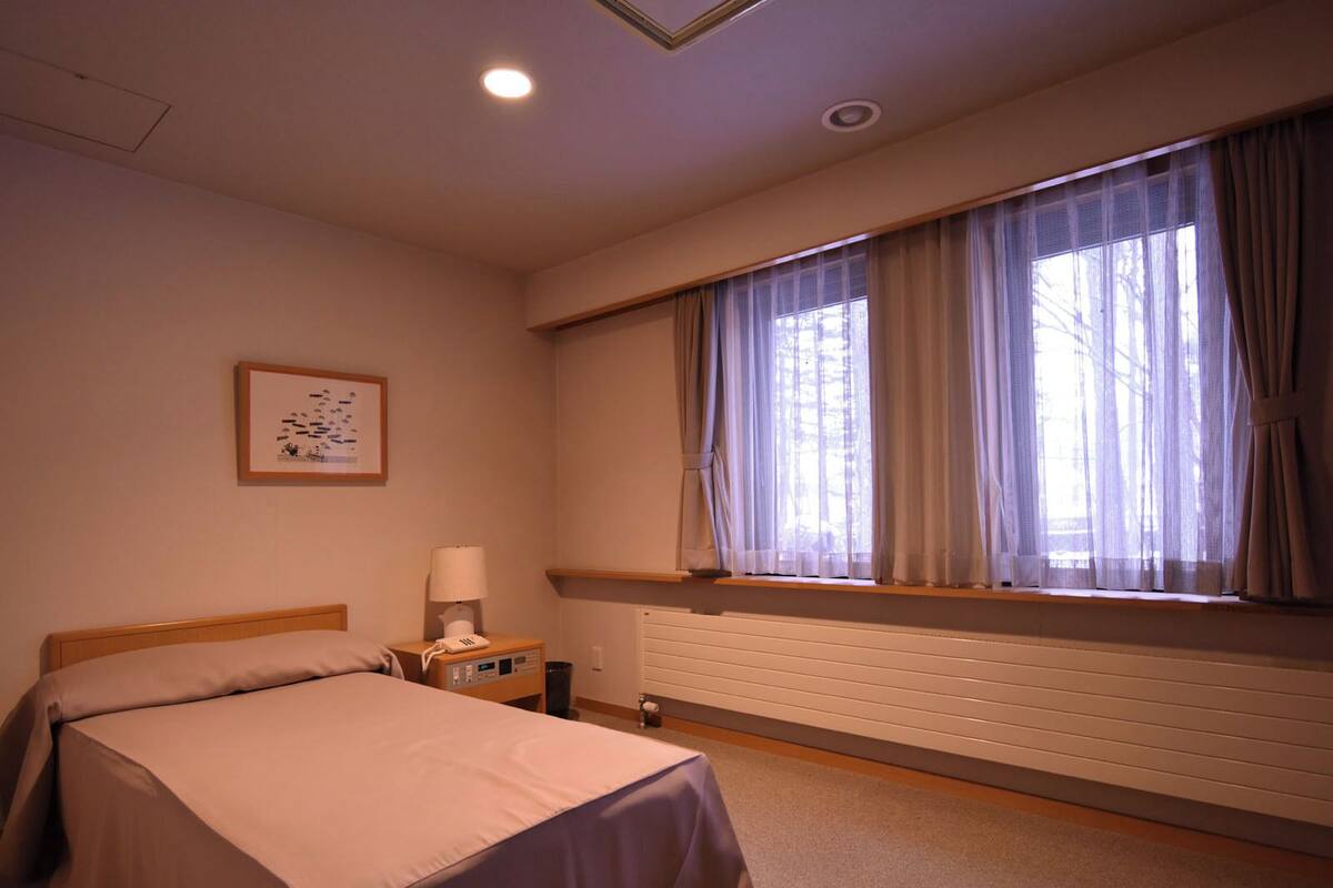A hotel room with a bed, a nightstand, a lamp, a window with curtains, and a radiator.