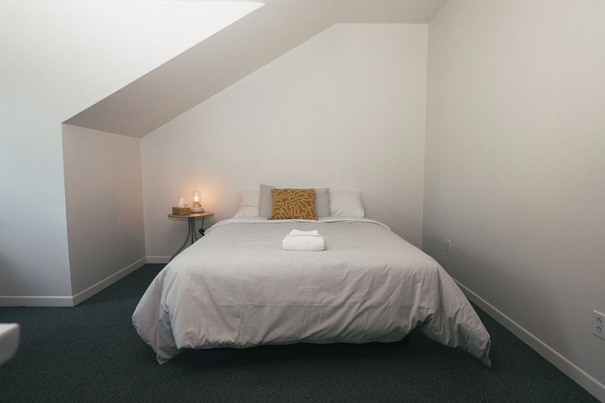 A neatly made bed with a white and gray striped bedspread, a white pillow, and a brown patterned pillow. A small bedside table with a lamp is visible to the left of the bed.
