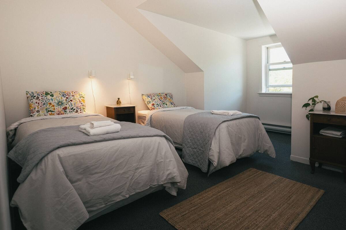 A bedroom with two beds, a nightstand, a dresser, a window, and a ceiling with a sloped design.