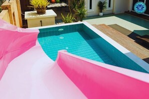 Una piscina con un tobogán rosado, rodeada de plantas en macetas y un símbolo de loto.