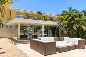A modern outdoor seating area with a glass enclosure, a large cushioned sofa, and a wooden deck. - Steps to Beach and 5th Ave, Resort Amenities (Playa del Carmen)