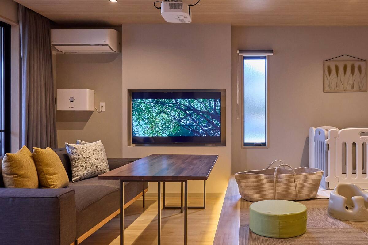 A modern living room with a flat-screen TV, a sofa, a table, a baby play area, and a window.