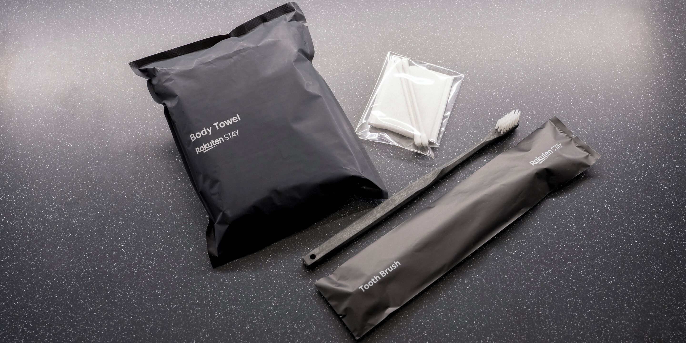 A black package labeled "Body Towel" and a separate package labeled "Tooth Brush" on a speckled surface.
