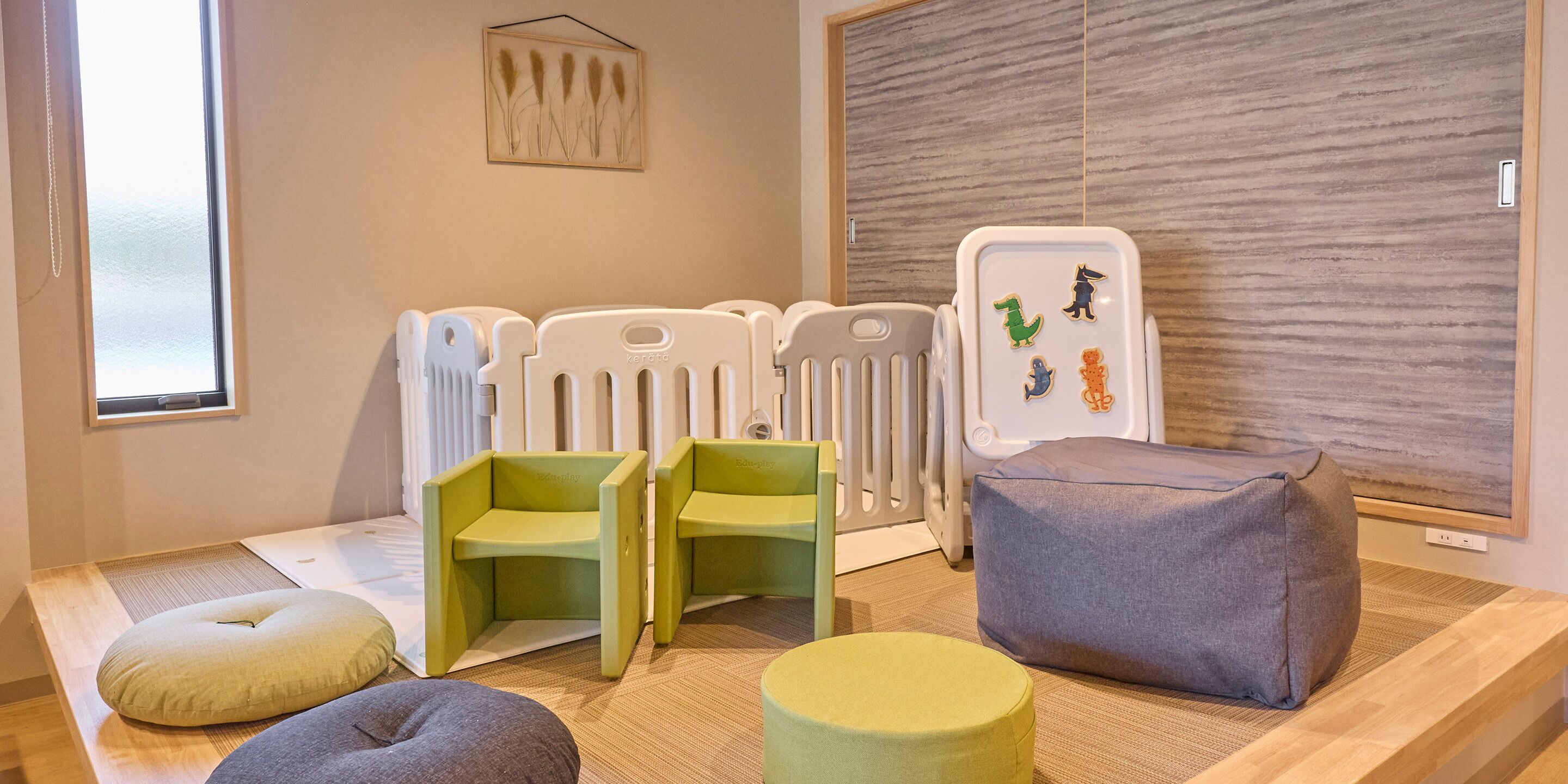 A room with a wooden floor, a window, a crib, two green chairs, a grey cushion, and a wall-mounted artwork.