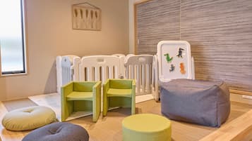 A room with a wooden floor, a window, a crib, two green chairs, a grey cushion, and a wall-mounted artwork.