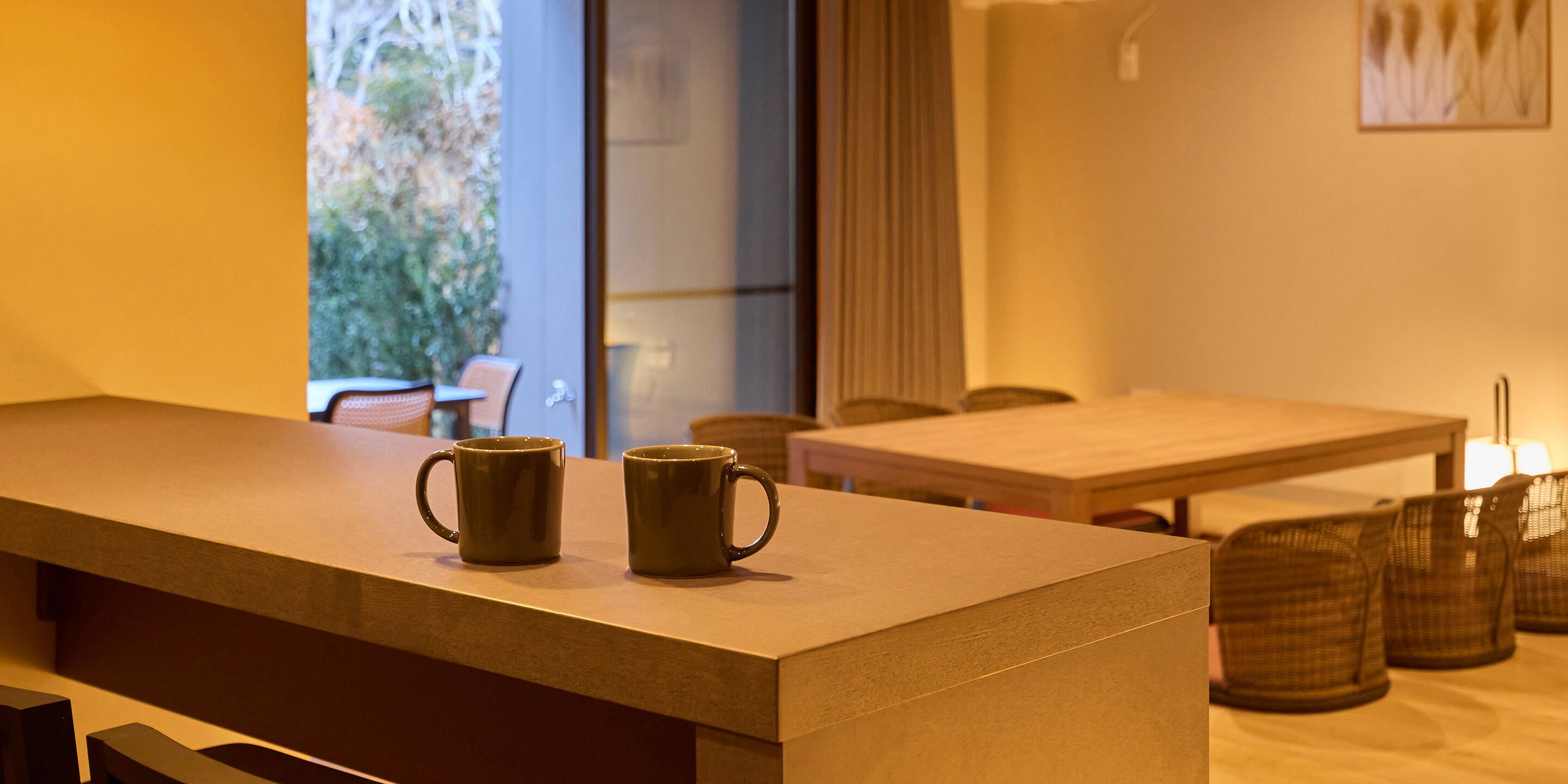 A reception area with a wooden counter and two green mugs. There is a table and chairs in the background, and a window with a view of greenery.