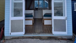 Una cabaña pequeña de azul claro con el número 447 en el techo, que cuenta con una zona de cocina, dos áreas de descanso con bancos y un ventanal con vistas a la playa.