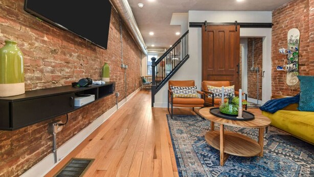 Un salon moderne avec un mur en briques, un téléviseur à écran plat, un plancher en bois, un escalier et un canapé jaune.