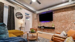 Un salon moderne avec un mur en briques, un téléviseur à écran plat fixé au-dessus d’une étagère, et un coin salon confortable agrémenté d’un tapis à motifs.